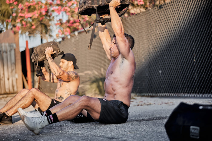 Two men performing outdoor rucking exercises with GORUCK sandbags under sunlight