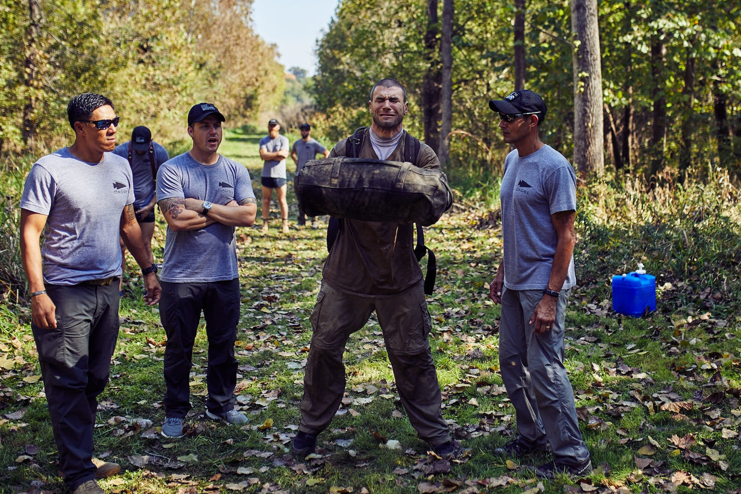 GORUCK team outdoors, man lifting heavy sandbag, group in athletic rucking gear training