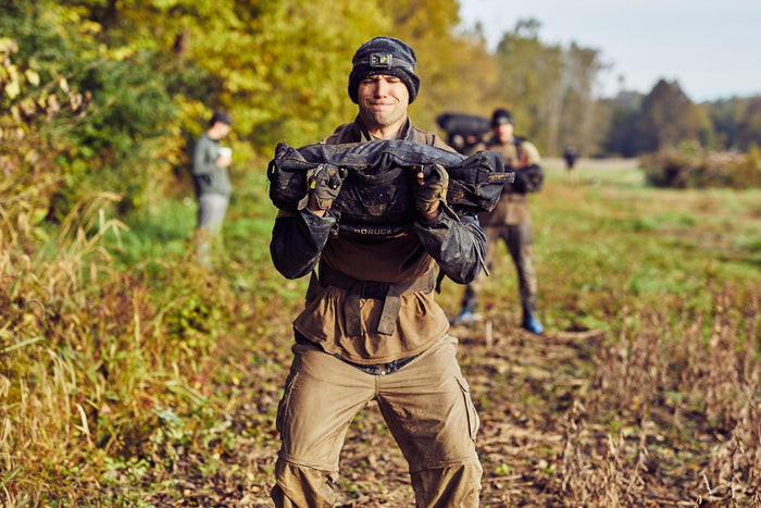Man lifting GORUCK training sandbag outdoors, rucking gear in muddy field