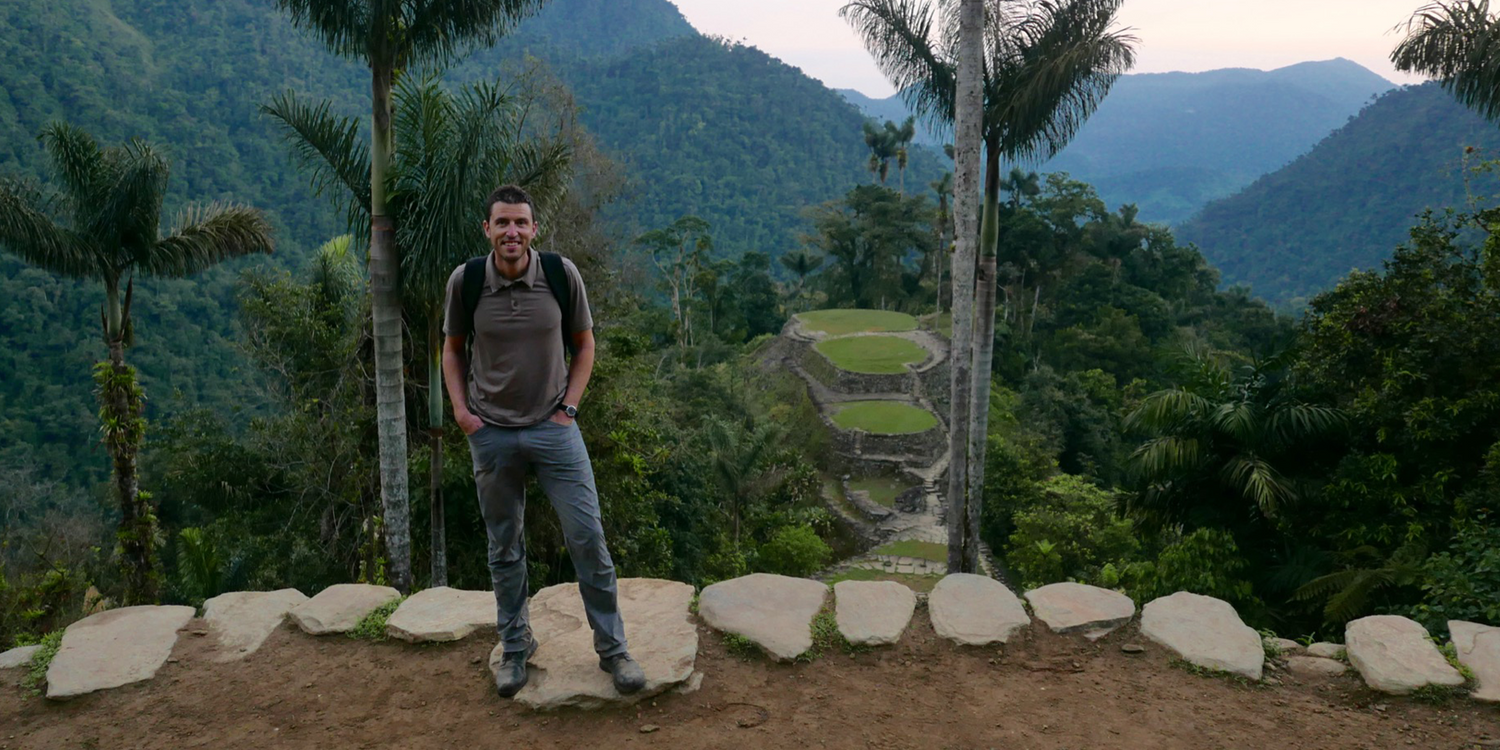 Man wearing GORUCK gear standing on stone path with lush mountains and jungle ruins behind