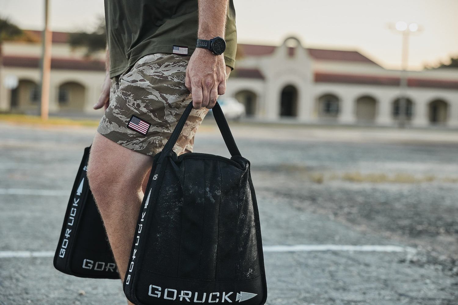 Man carrying GORUCK sandbags in outdoor training area, wearing camo shorts with US flag patch.