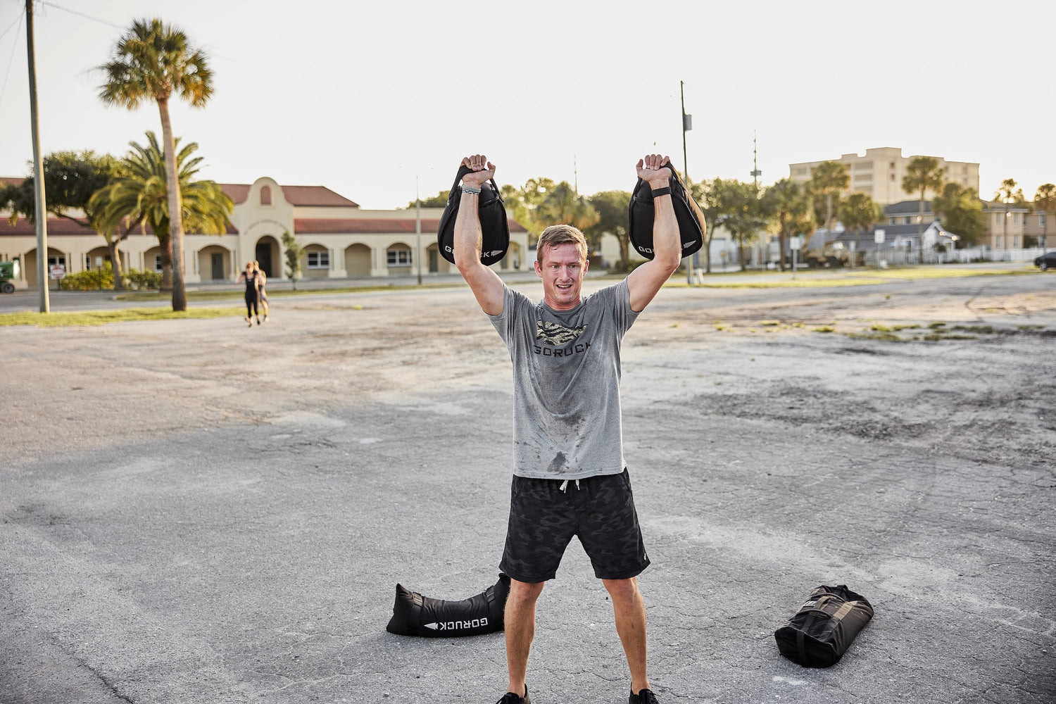 Man outdoors lifting GORUCK sandbags overhead, wearing GORUCK shirt, athletic training scene