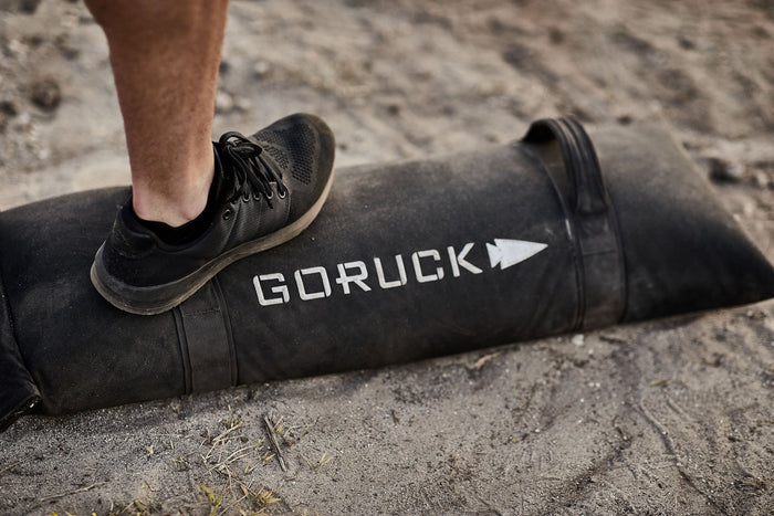 GORUCK sandbag with logo outdoors, person wearing black rucking shoe on top