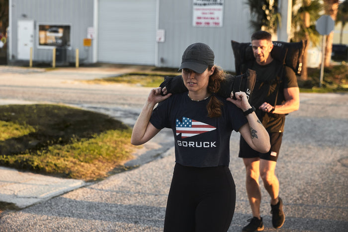 Two people rucking outdoors with weighted gear, woman wearing GORUCK shirt featuring flag logo