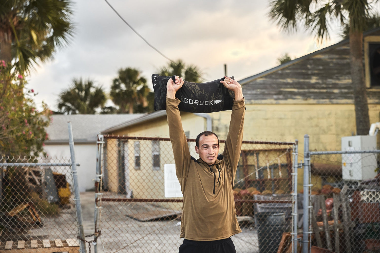 Man lifting GORUCK sandbag overhead outdoors, wearing athletic gear, showcasing rucking fitness