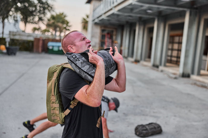 Man training outdoors with GORUCK sandbag and rucksack, demonstrating tough rucking gear