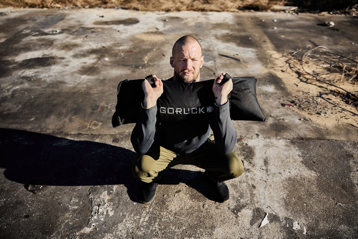 Man squatting outdoors with GORUCK sandbag, demonstrating rugged rucking training gear.