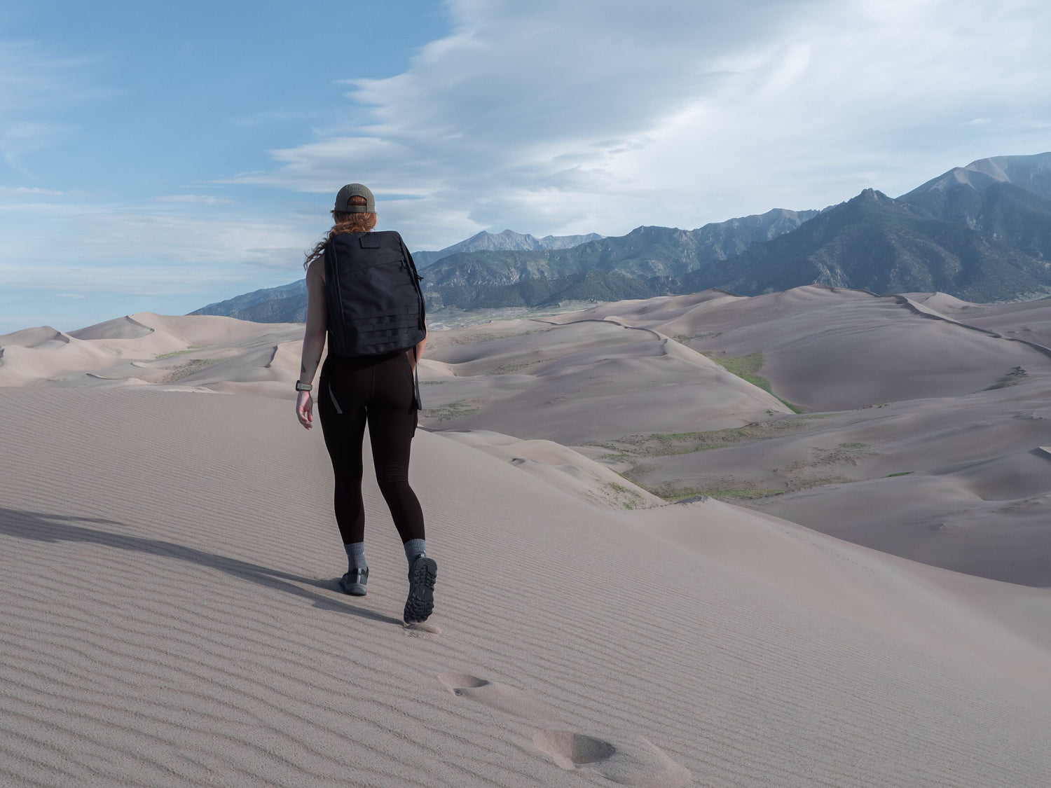 Woman rucking with GORUCK backpack on sand dunes, mountains in background under blue sky