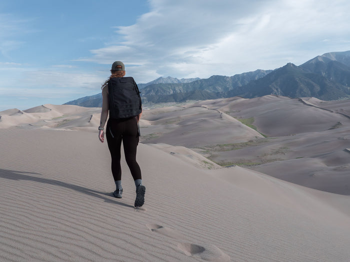 Woman rucking with GORUCK backpack on sand dunes, mountains in background under blue sky