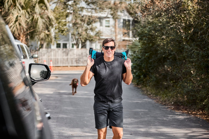 Man rucking outdoors with GORUCK sandbag, wearing black shirt and shorts, dog in background