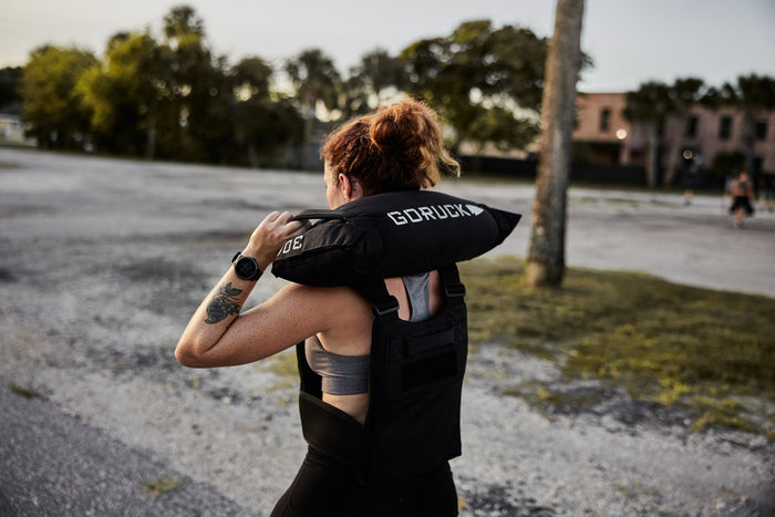 Woman wearing GORUCK vest and carrying training sandbag outdoors during rucking workout