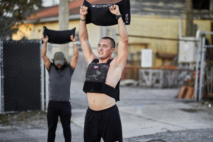 Man wearing GORUCK weighted vest lifting sandbag overhead during outdoor rucking workout