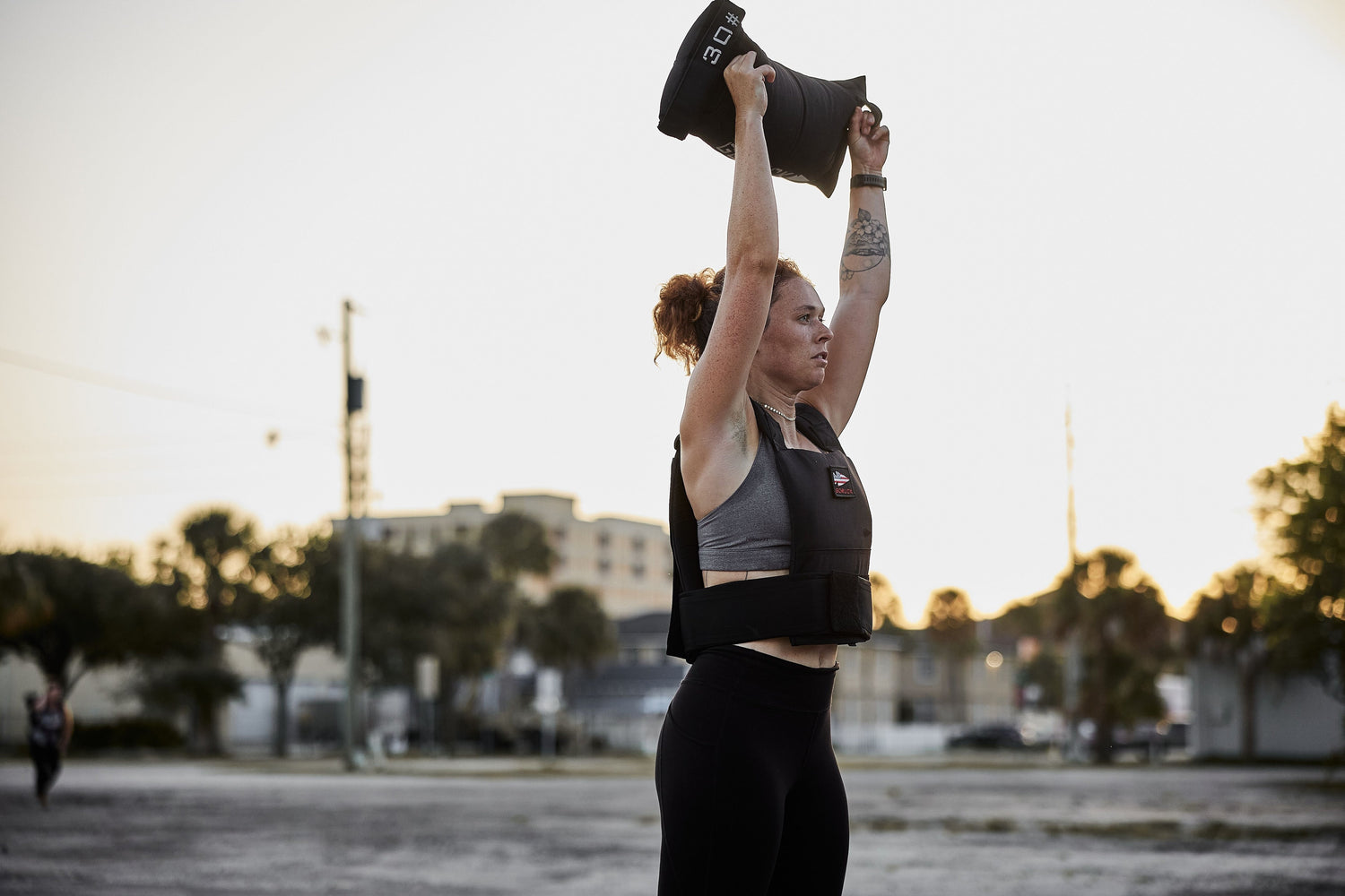 Woman outdoors lifting weighted sandbag overhead wearing GORUCK ruck plate carrier vest