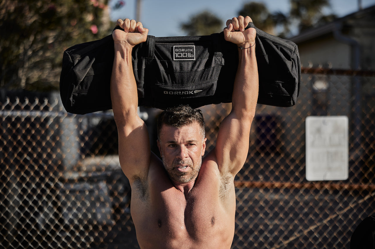 Man performing outdoor strict press with GORUCK sandbag, showcasing rucking gear strength