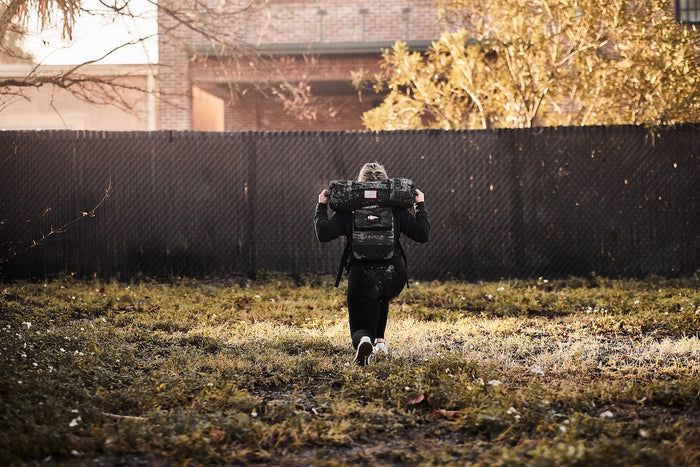 Person rucking outdoors with GORUCK backpack and sandbag, training in grassy field