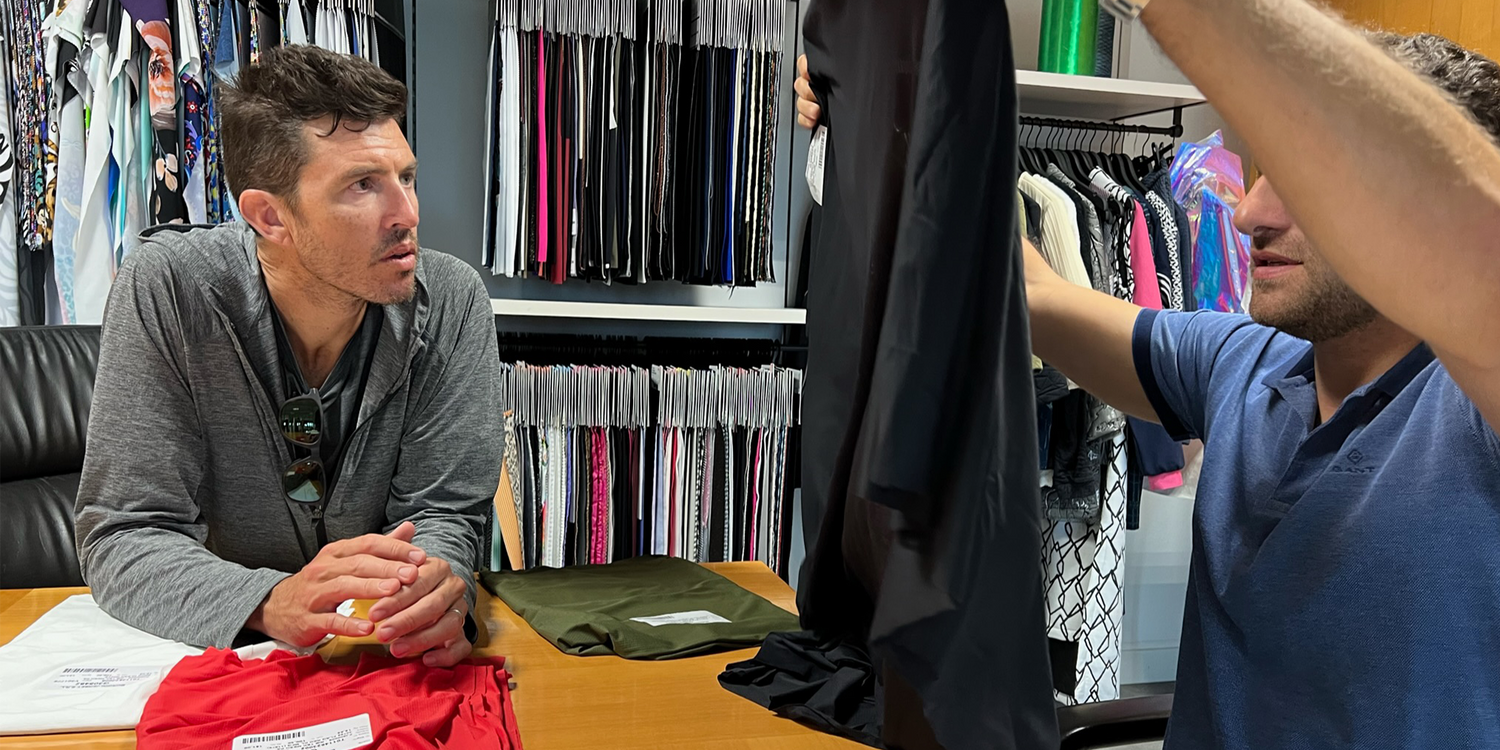 Two men examining and discussing black fabric samples in a textile showroom with colorful fabric swatches on shelves