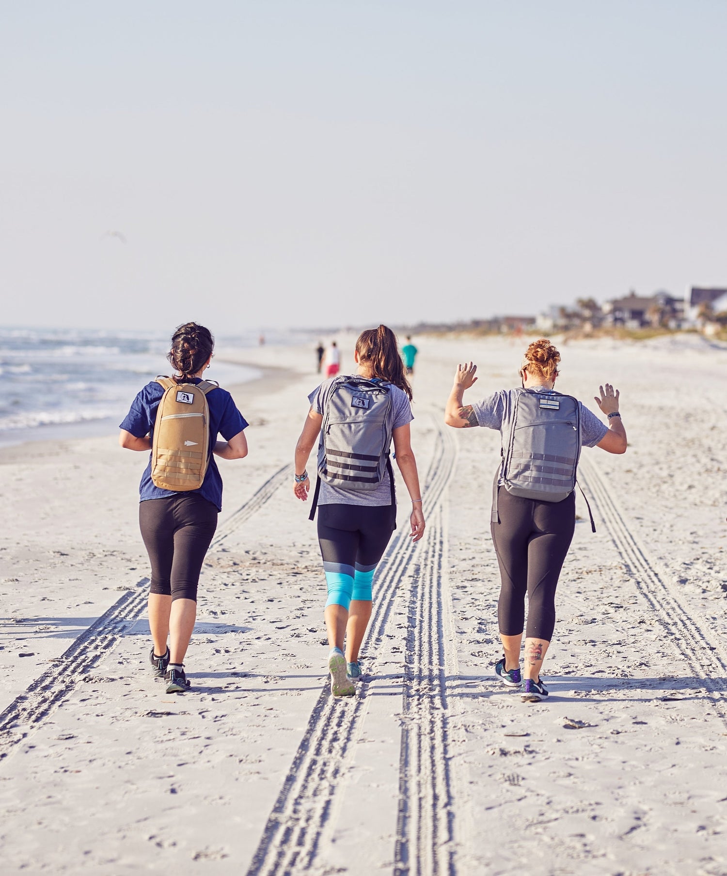 Three women rucking on a sandy beach with GORUCK backpacks, ocean and homes in background