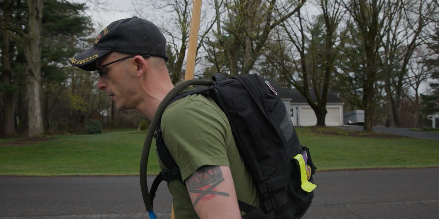 Man in black cap and green shirt rucking with GORUCK backpack and hydration tube on residential street