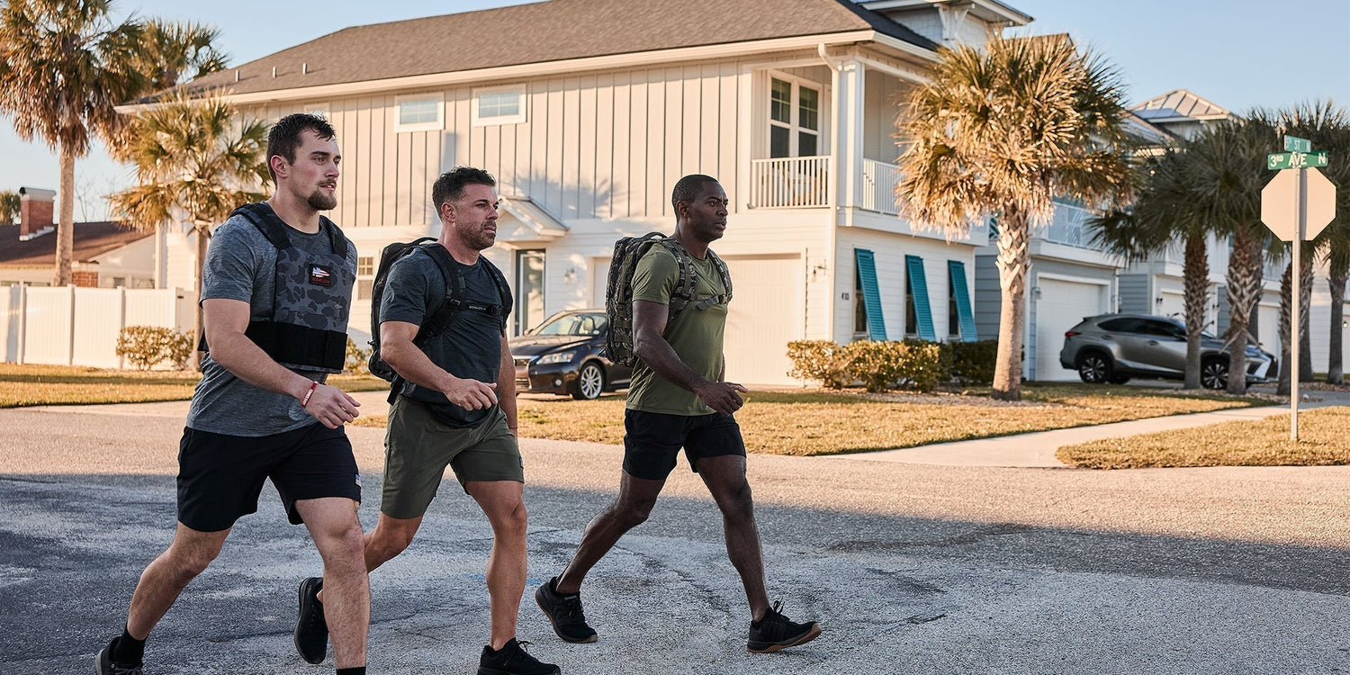 Three men in athletic gear wearing weighted vests and backpacks walking in a sunny residential neighborhood with palm trees