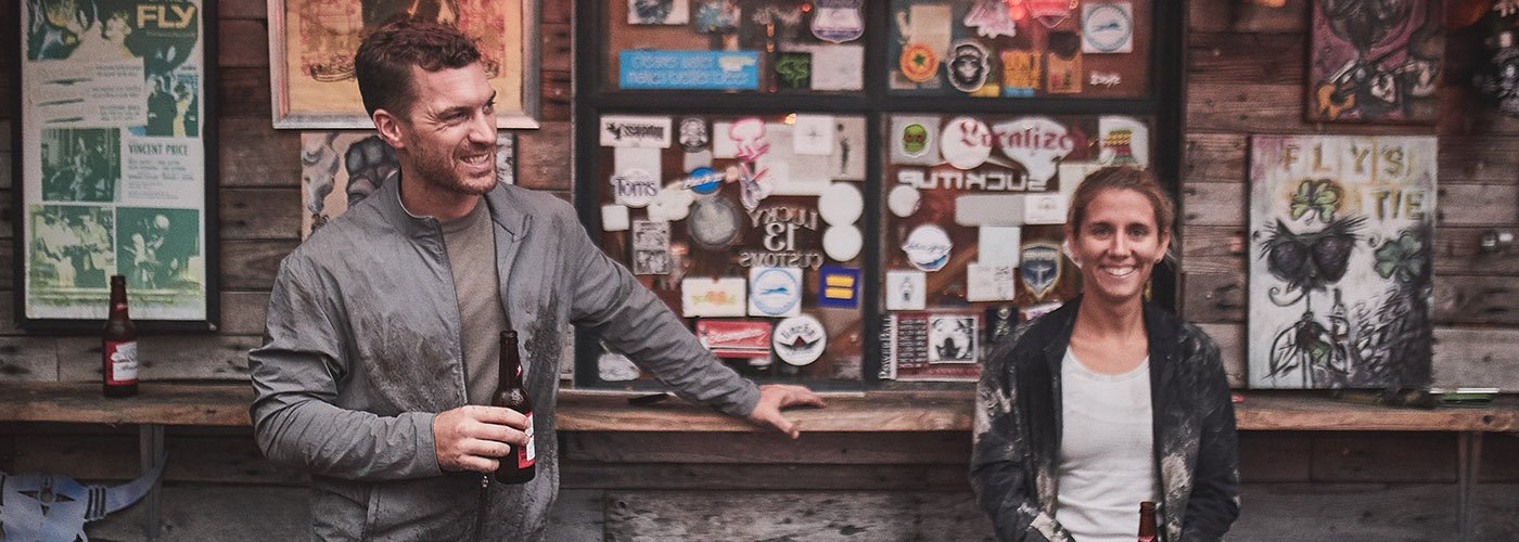 Two people holding beer bottles smile and chat at a bar with posters and stickers on the wooden wall behind them.