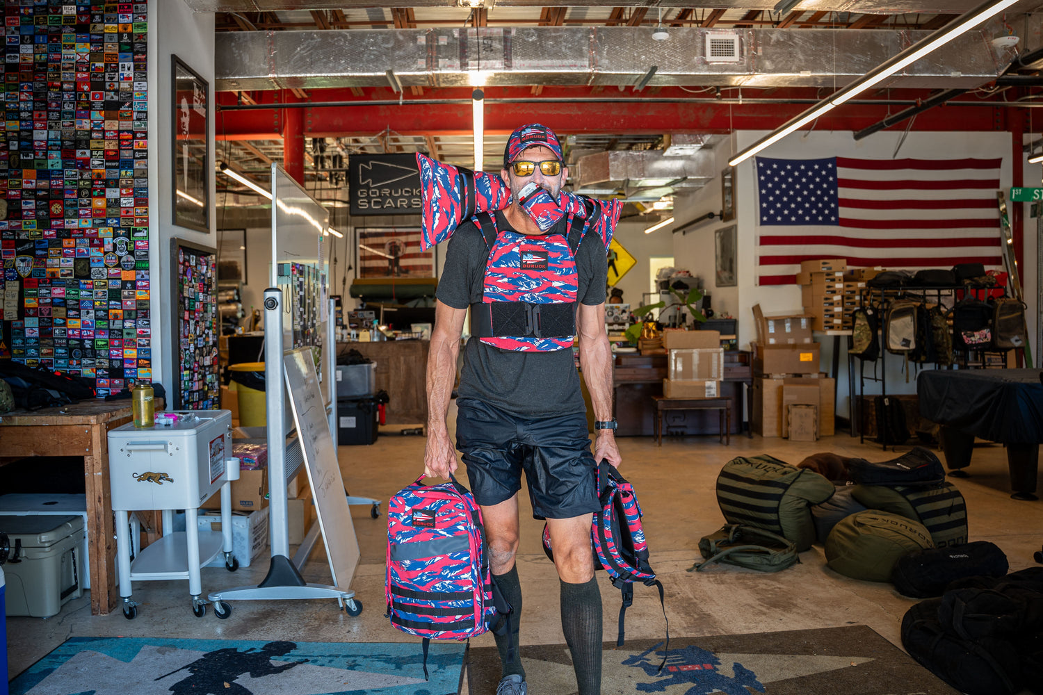 A man in a workshop wears matching colorful backpacks on his back, chest, shoulders, and holds one in his hand.