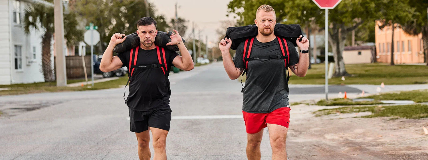 Two men walking on a street carrying weighted rucking bags with red straps, wearing athletic gear