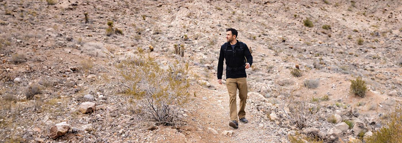 Man wearing GORUCK rugged gear walking on rocky desert trail with dry bushes