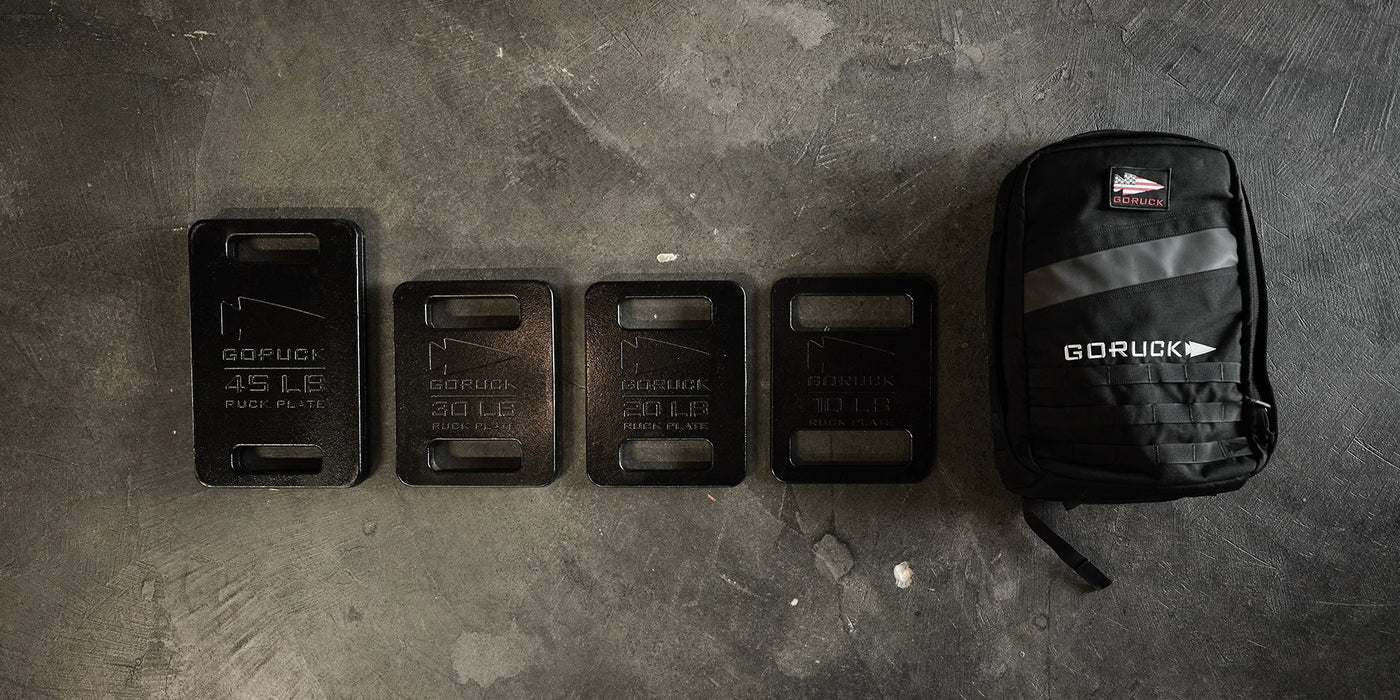 Four black weight plates of different sizes lined up next to a black GORUCK backpack on a concrete floor.