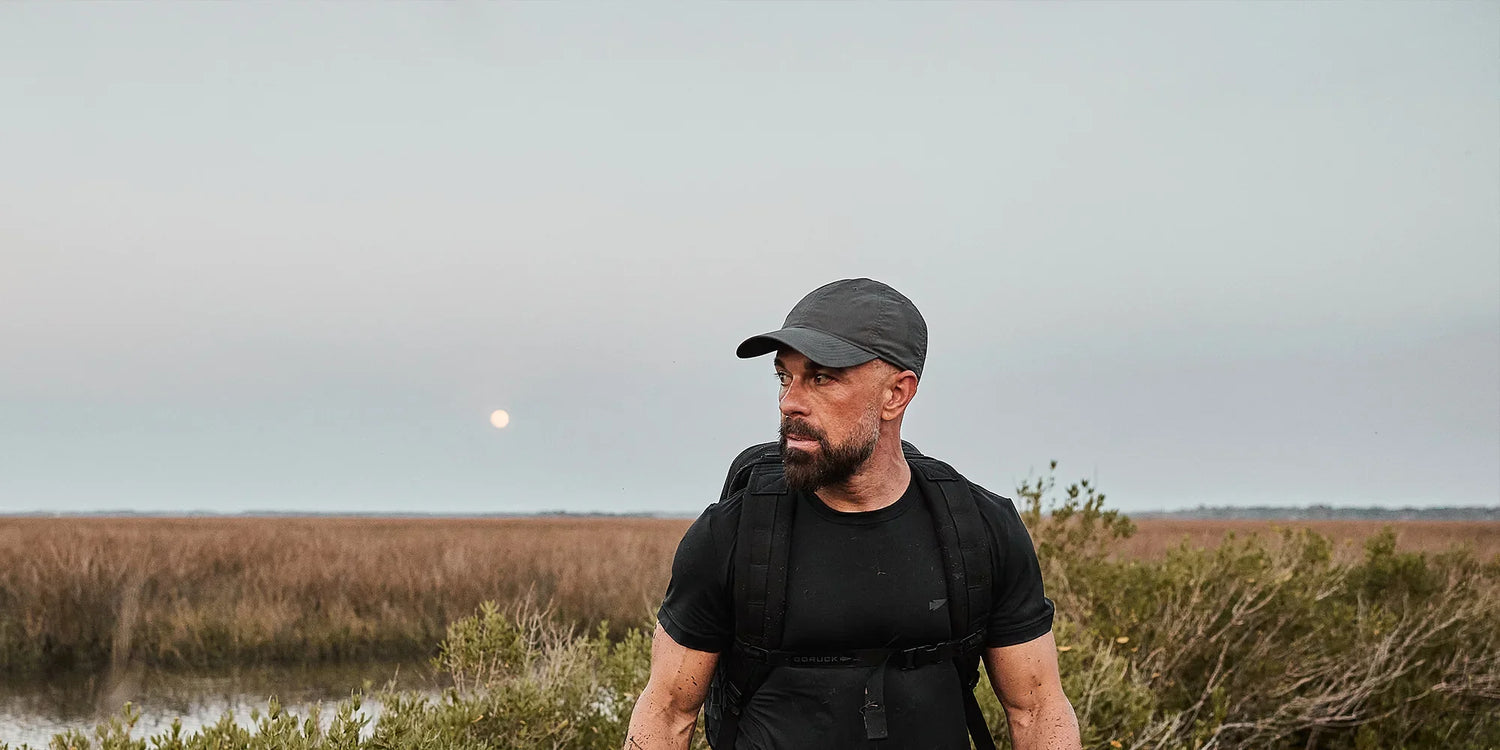 Man wearing black GORUCK gear and cap standing in outdoor marshland during early evening