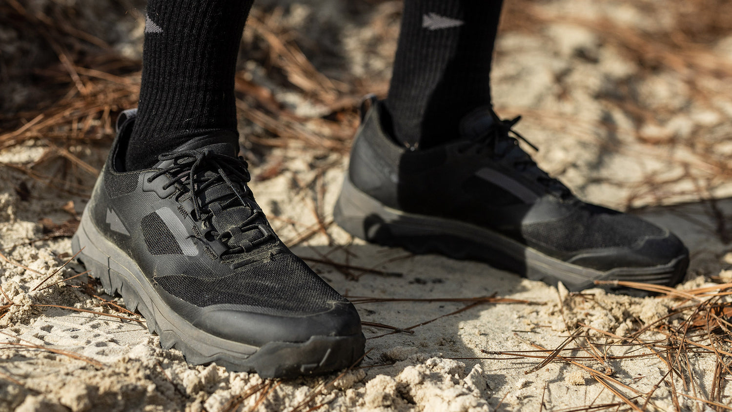 Close-up of a person wearing black hiking shoes with black socks, standing on a sandy ground covered with dry pine needles.