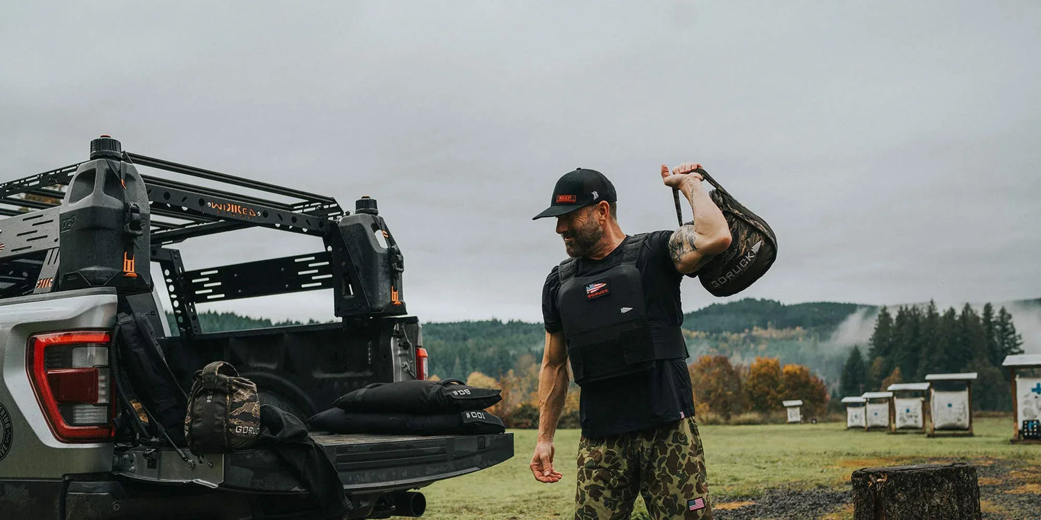 Man wearing tactical vest and camo pants holding GORUCK weighted bag near truck with outdoor gear in forested field