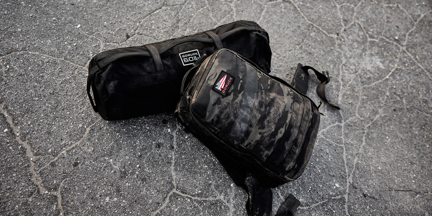 Two black duffle bags are placed on a cracked asphalt surface. One bag is labeled GORUCK 30L and the other has a camouflage pattern. Both bags are visibly worn and used.