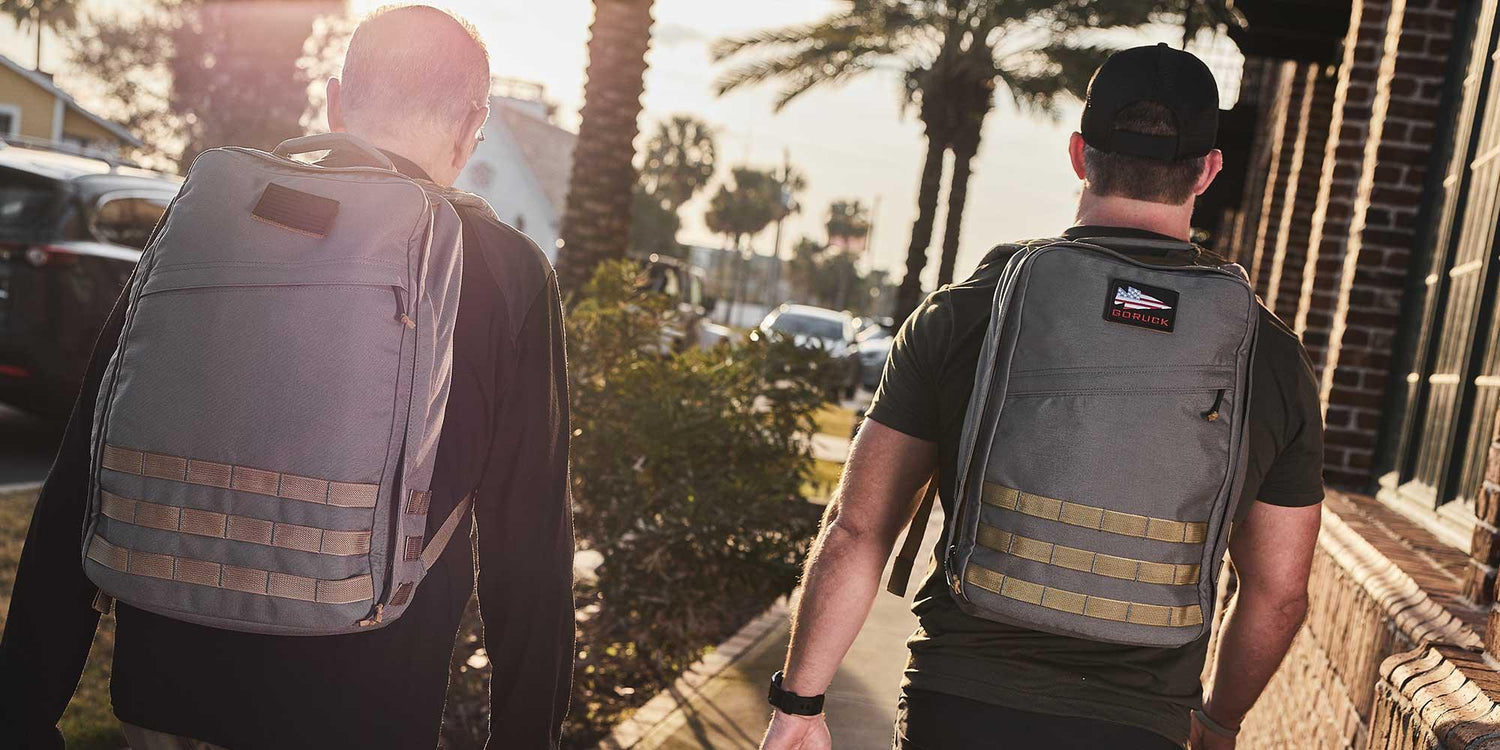 Two men walking outdoors in sunlight wearing durable grey GORUCK backpacks on a palm tree-lined street
