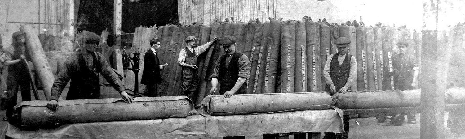 Workers rolling large carpets in a factory setting, surrounded by stacked materials.