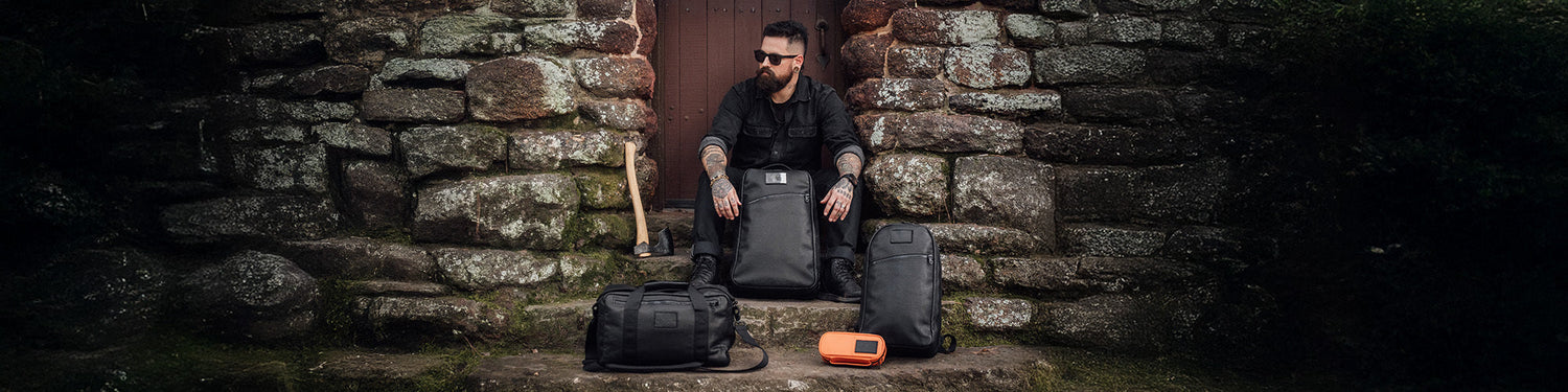 A man in black sits on stone steps with three black bags, an orange pouch, and an axe nearby.