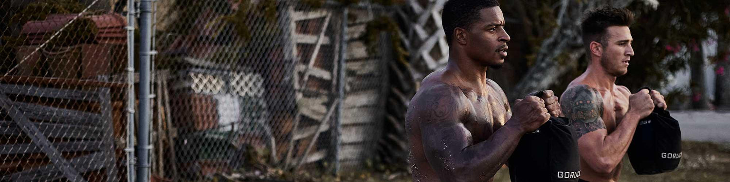 Two muscular men doing kettlebell exercises outdoors holding black GORUCK sandbags