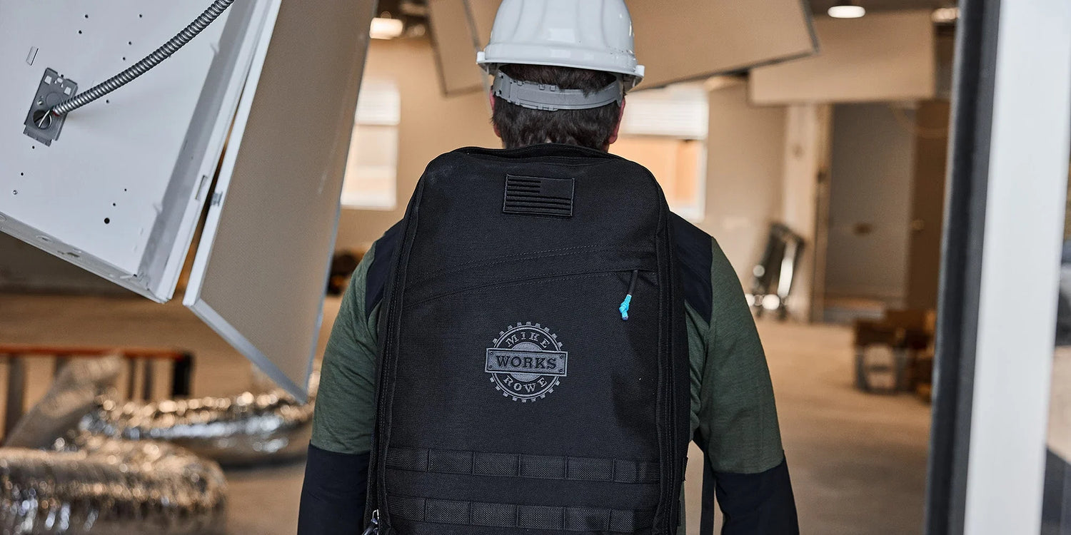 Man wearing a white helmet and black Mike Rowe Works backpack inside a construction site with exposed ducts