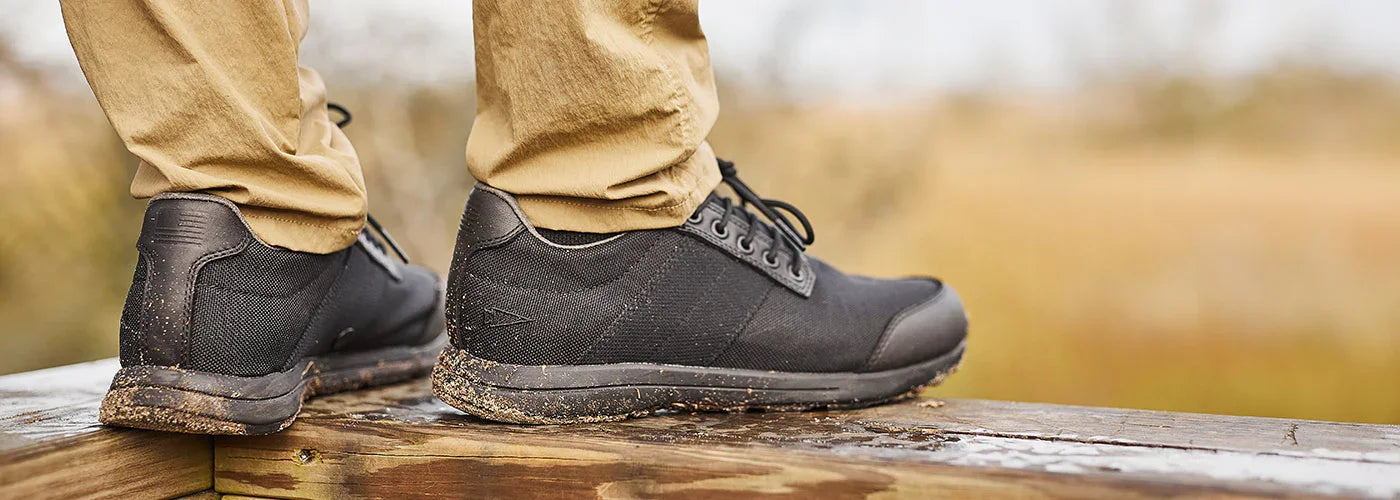 Close-up of black GORUCK cross trainers with mud on soles, worn with tan pants on wooden surface outdoors