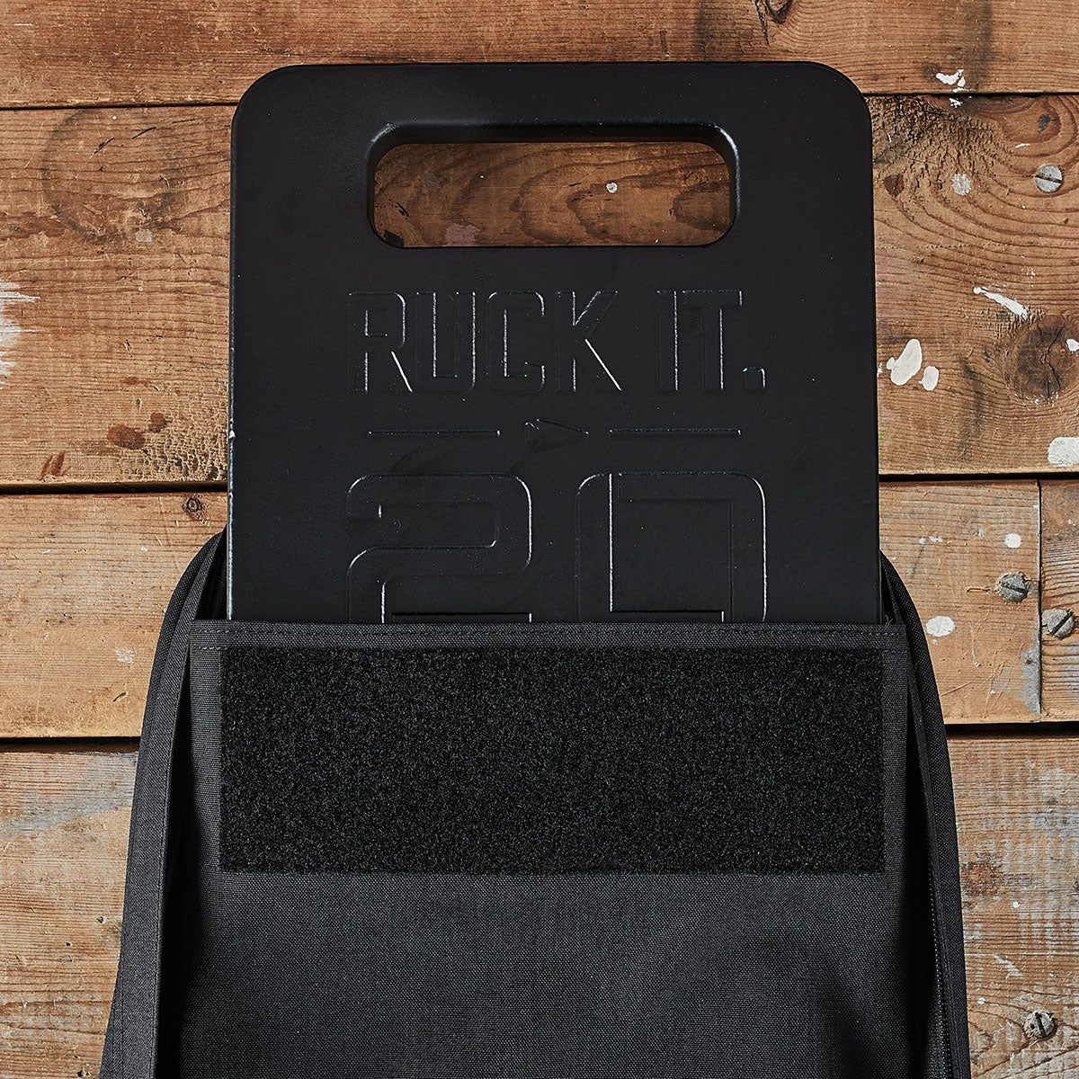 A black weight plate labeled "Ruck It. 20" in a black backpack against a wooden background.