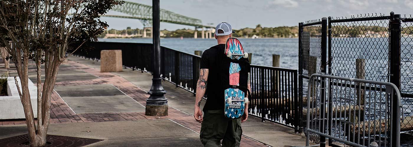 Man carrying colorful skateboard walking on riverside pathway with bridge and waterfront background