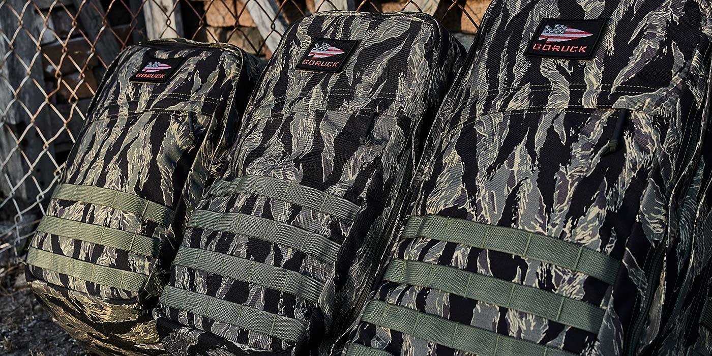 Three GORUCK branded tactical rucking backpacks in tiger stripe camouflage pattern leaning against a chain-link fence