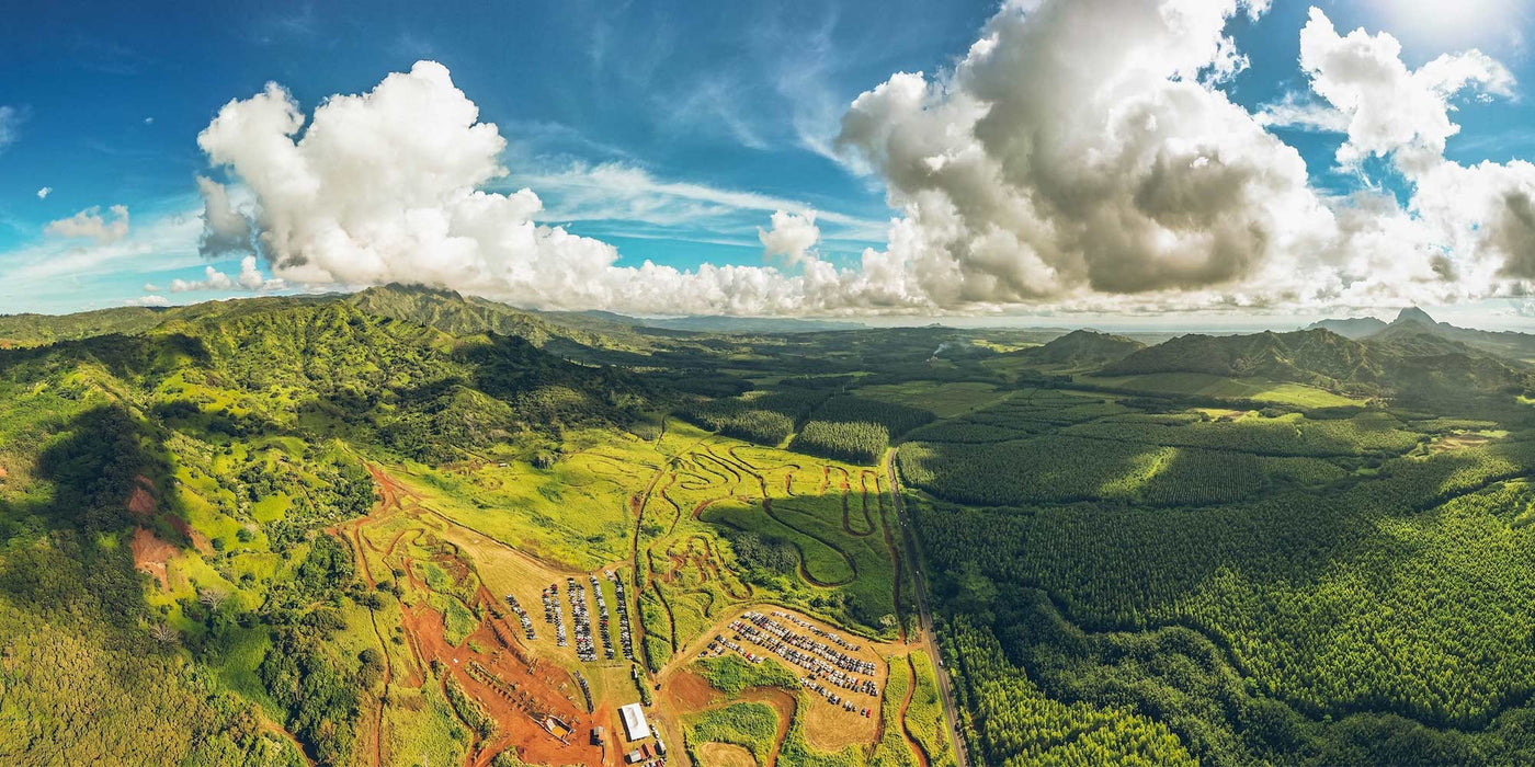 Aerial view of a lush green landscape with mountains, forests, and a small settlement under a blue sky with clouds.