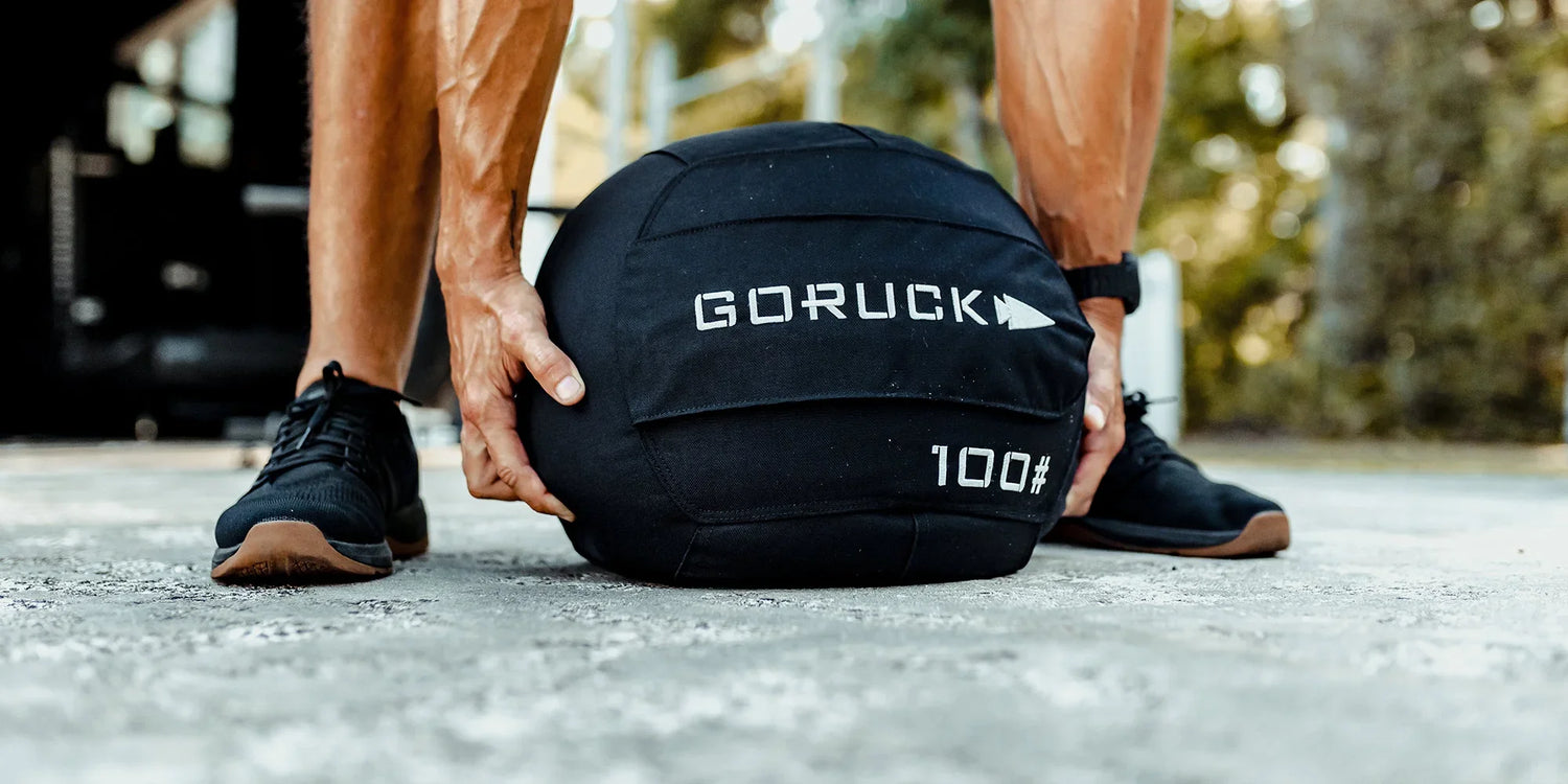 Person gripping a black GORUCK 100-pound weighted sandbag on concrete outdoors for strength training