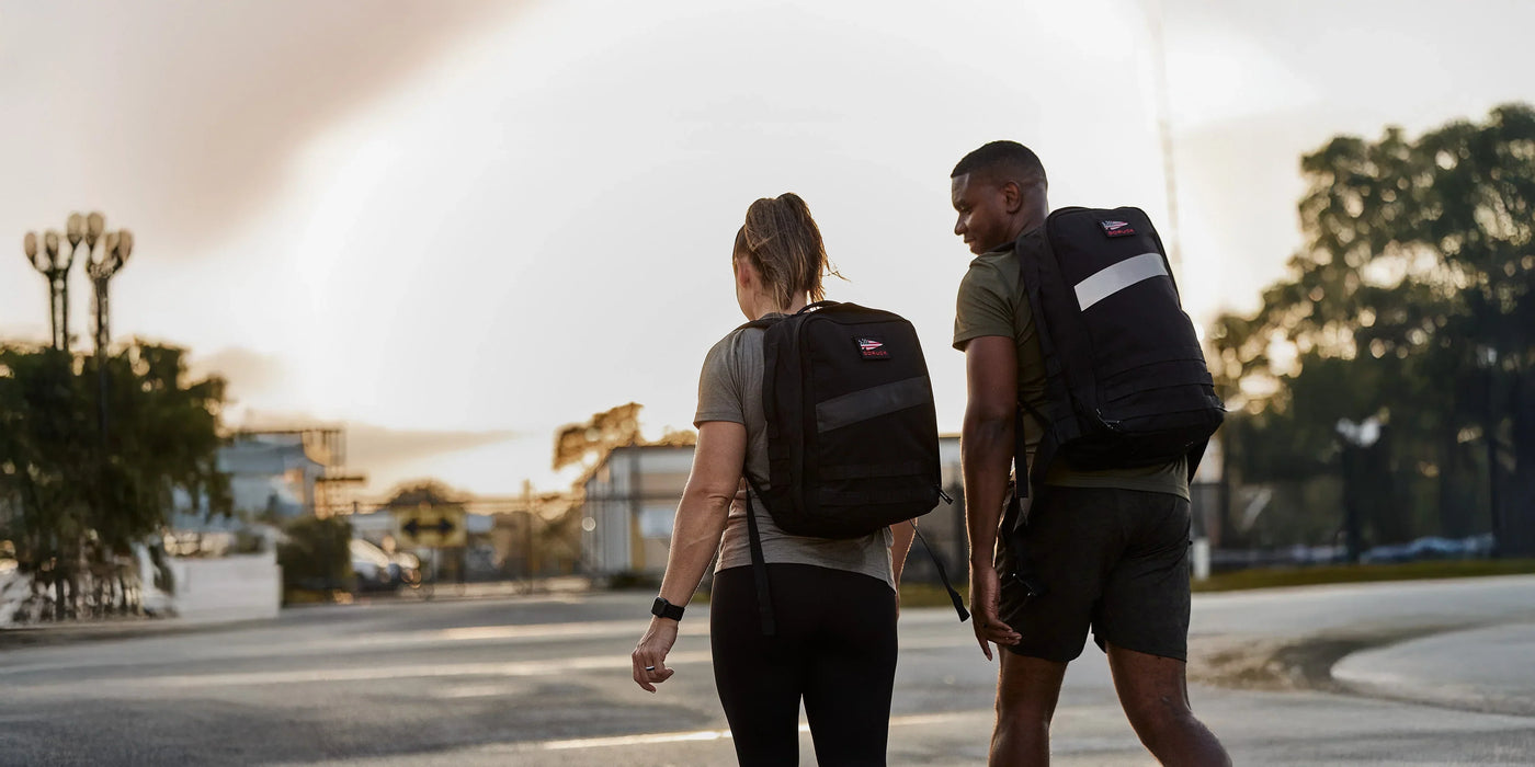 Two people with backpacks walk outside on a sunny day, wearing athletic clothes and facing away from the camera.