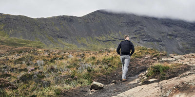 A person with a backpack hikes up a rocky trail toward a mountain under a cloudy sky.
