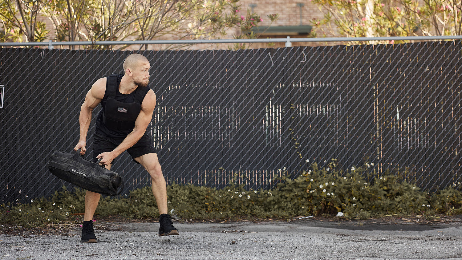 A man in athletic wear lifts a heavy bag outdoors near a chain-link fence and some bushes.