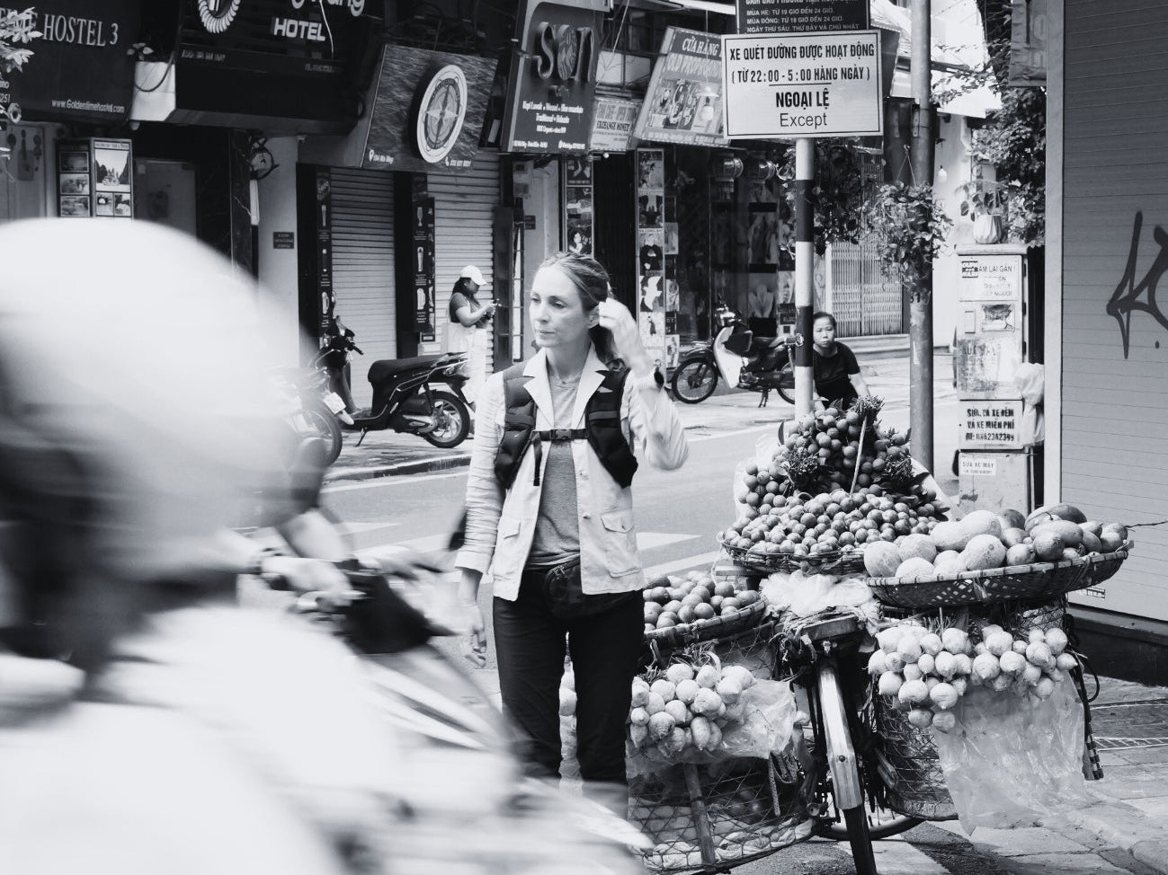 Woman stands by a fruit-laden bicycle on a busy street with blurred motorbikes passing by.
