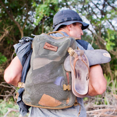 A man walks outdoors near trees, wearing a hat and the Mesh Ruck backpack with sandals attached.