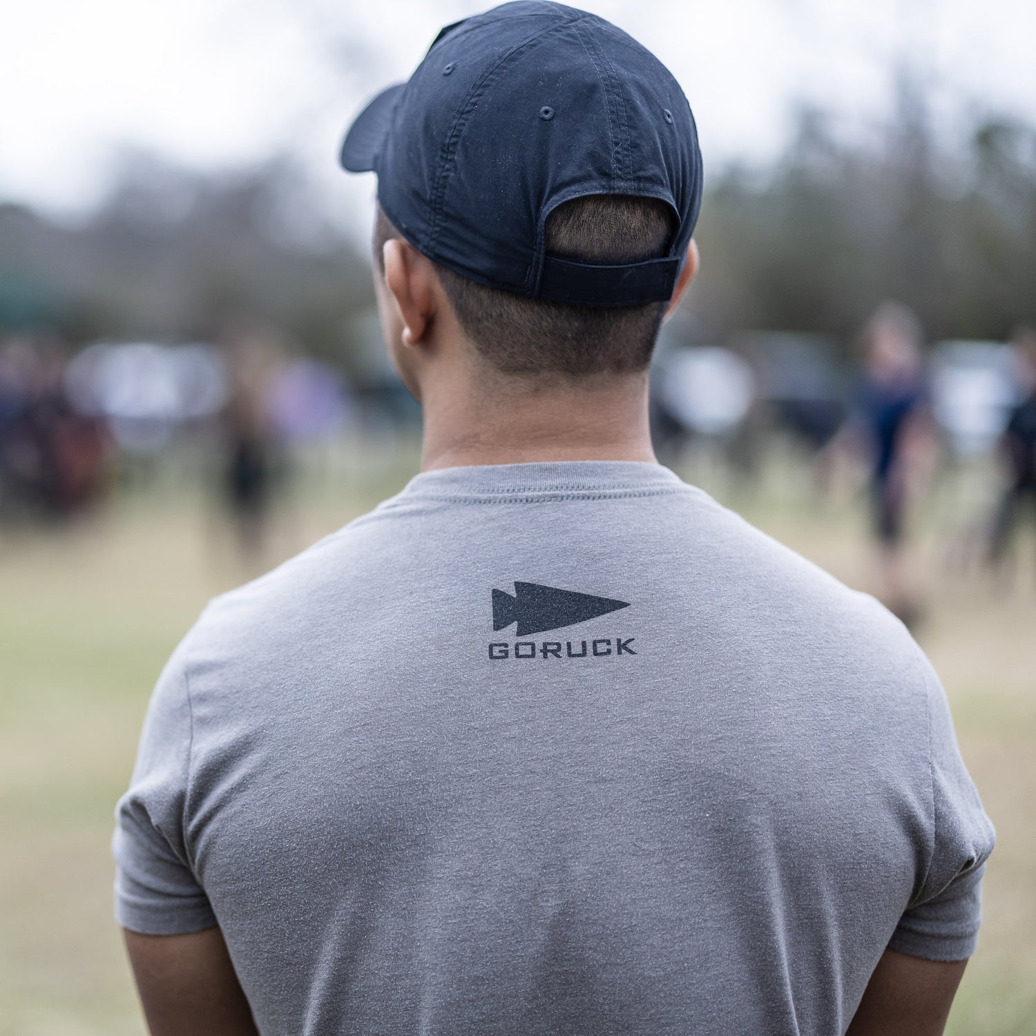 A man wearing the Subtle Spearhead Tee - Unisex Tri-Blend in gray and a navy cap stands outdoors, facing away, with blurred people visible in the background.