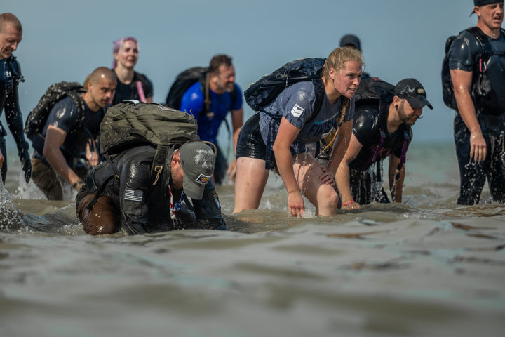 A group of people with backpacks wade through shallow water, appearing tired but determined.